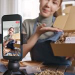 A woman films an unboxing video in front of a recording phone.