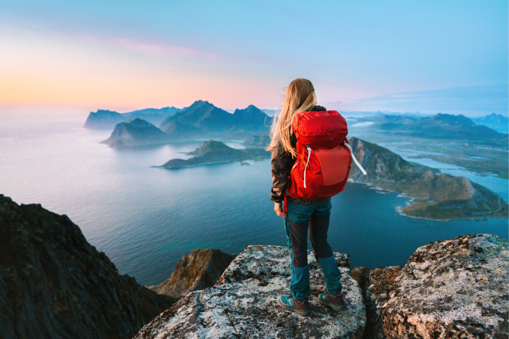 A woman stands on top of a mountain looking out at a body of water.