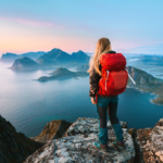 A woman stands on top of a mountain looking out at a body of water.