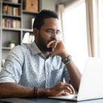 Focused young businessman sits at desk looking at laptop