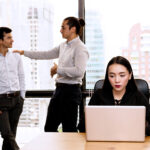 Woman uses her laptop as two male coworkers communicate behind her in an office
