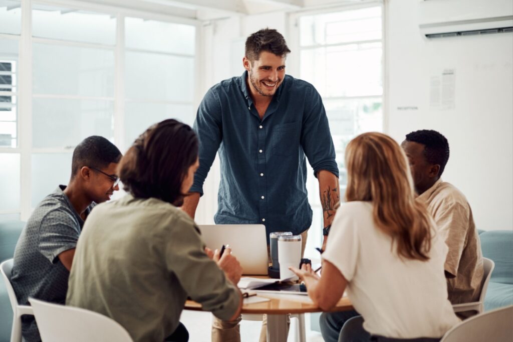 One man standing and a group of people sitting around a table.