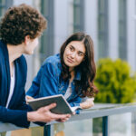 A woman attentively looks at and listens to a man, with a modern, urban building in the background.