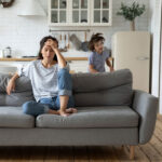 Exhausted mom sitting on couch with little children running playing