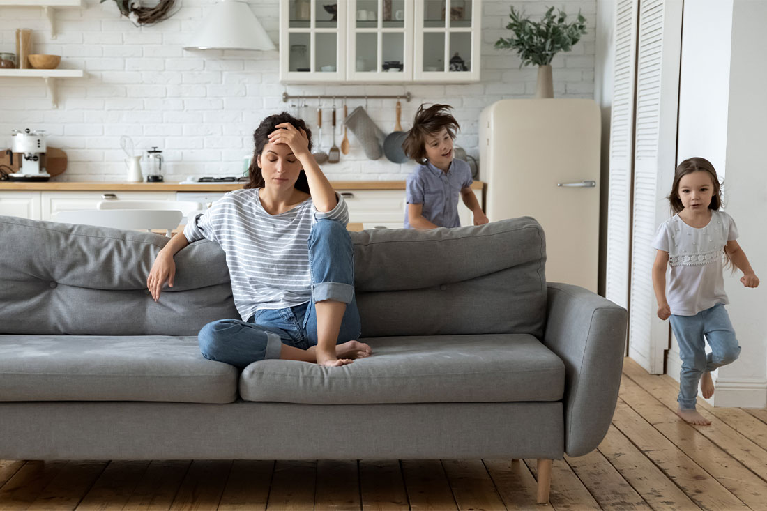 Exhausted mom sitting on couch with little children running playing