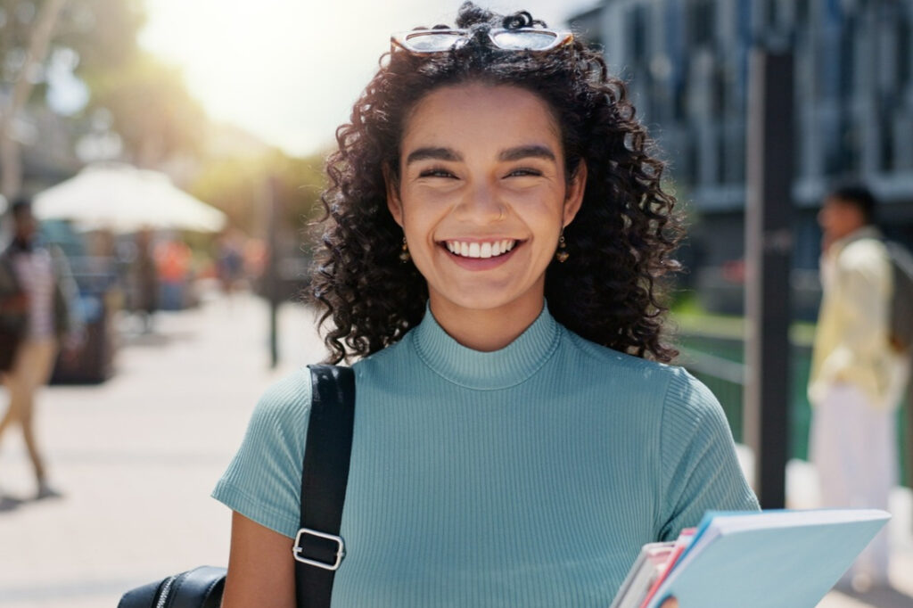 a college student smiling with a backpack hung over right shoulder and carrying notebooks in her left hand