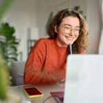 a woman sitting at her work desk while smiling at her laptop screen and holding a pen near her face