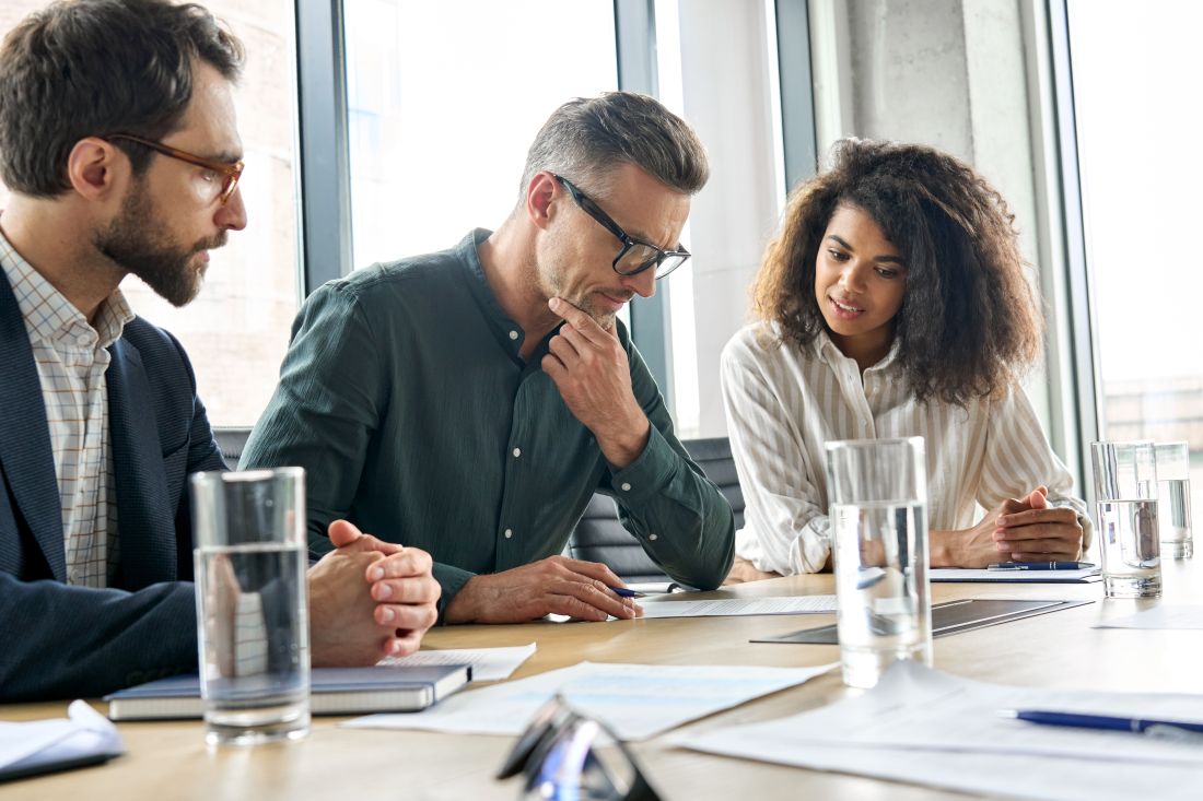 two businessmen and a businesswoman sitting at a desk in an office and reviewing a document