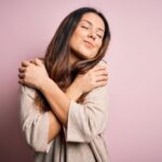 A woman hugging herself against a pink background.