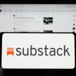 Person holding cellphone with logo of subscription company Substack on screen in front of business webpage