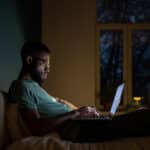 Focused man in dark room lying on bed work on laptop
