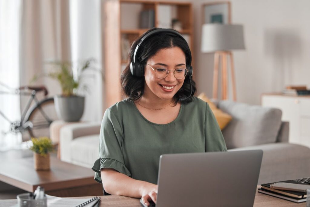a woman sitting at a desk in her home and wearing headphones while using her laptop
