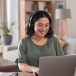 a woman sitting at a desk in her home and wearing headphones while using her laptop