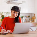 Asian woman writing notes in planner while taking online course at home using laptop computer