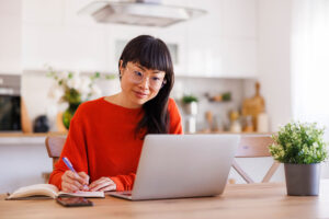 Asian woman writing notes in planner while taking online course at home using laptop computer