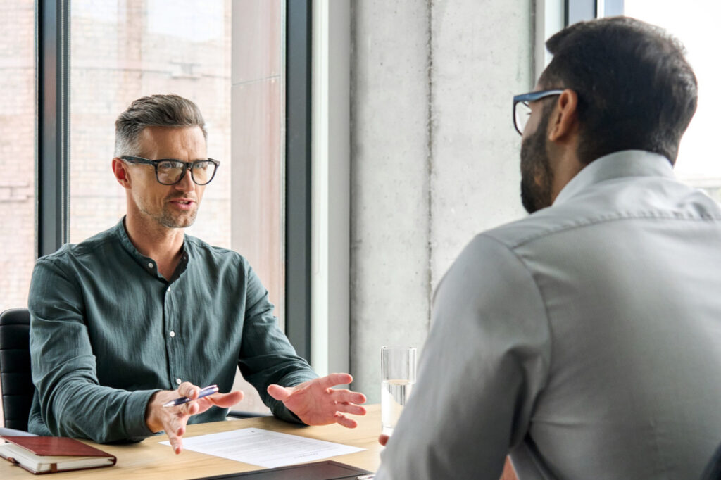 a boss sitting at a desk in an office and speaking to an employee
