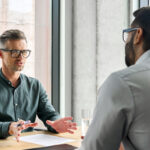 a boss sitting at a desk in an office and speaking to an employee