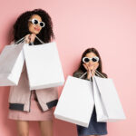 Fashionable mother and daughter in sunglasses holding shopping bags on pink background