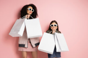 Fashionable mother and daughter in sunglasses holding shopping bags on pink background