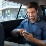 Happy smiling business man typing message on phone while sitting in a taxi.