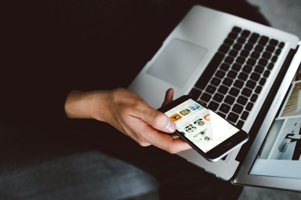 A person holds a smart phone in their hand and a laptop on their lap.