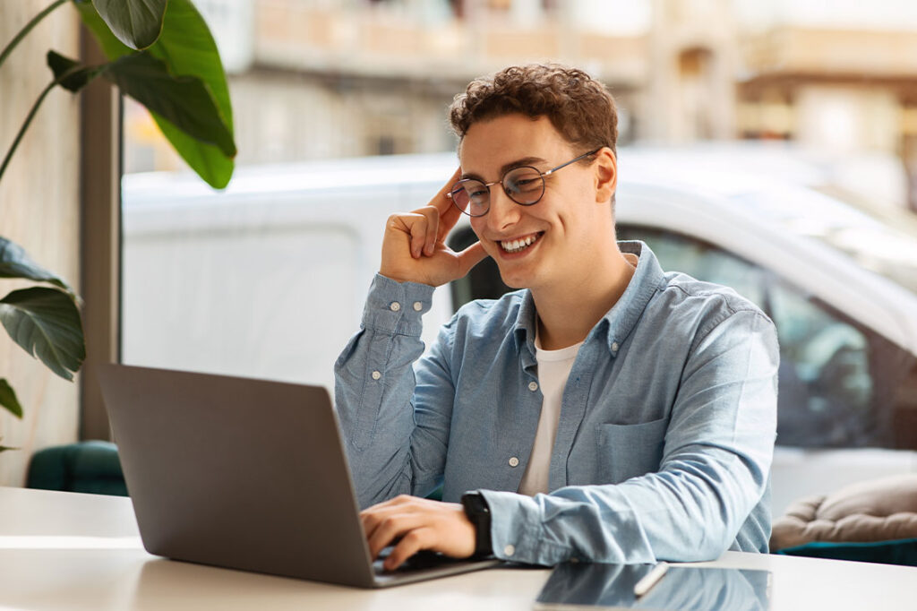 young man with curly hair and glasses smiling at his laptop in a bright room with a plant